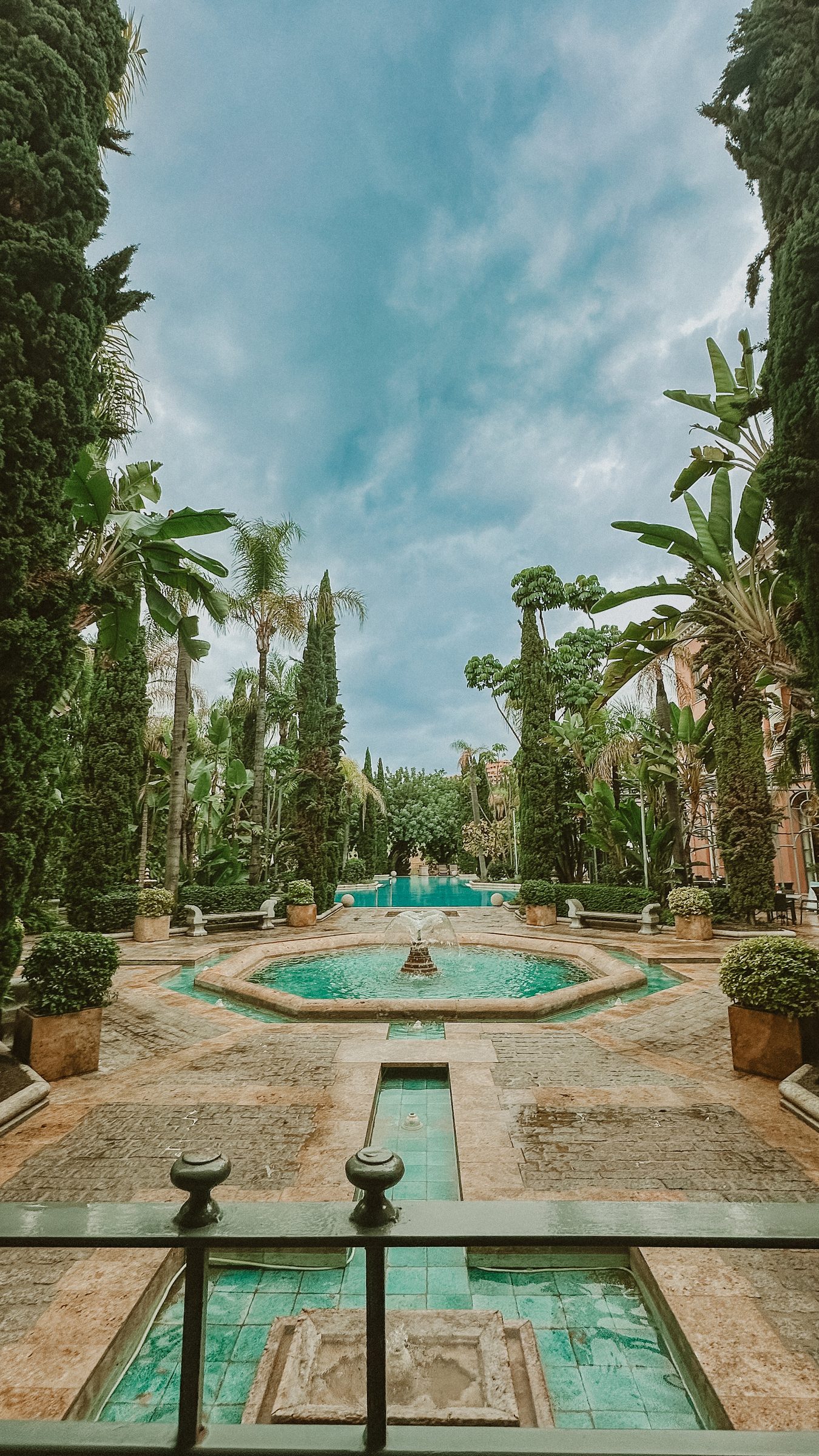 A formal garden with a stone water rill running between cypress and palm trees to a circular fountain