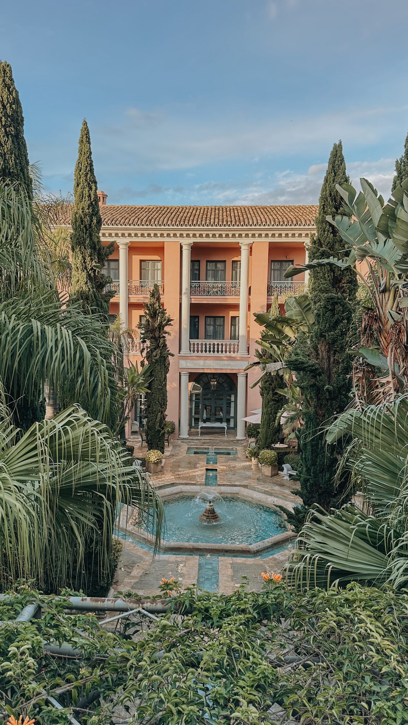 The terracotta palace facade seen from the garden, framed by palms and cypresses with a fountain in front