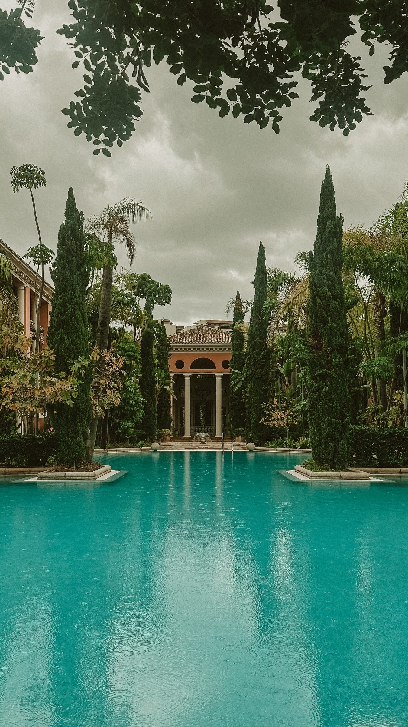 A long turquoise swimming pool lined with cypress trees leading to a columned stone pavilion