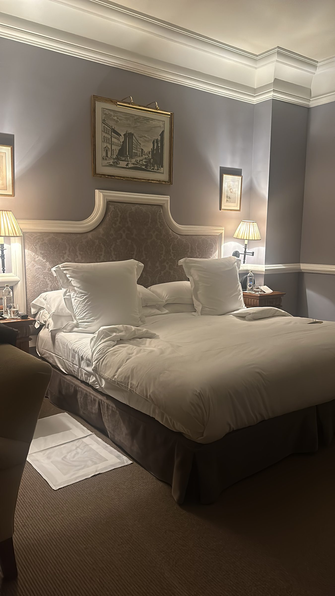 A hotel bed with white linen and an upholstered headboard, framed prints on the wall and lamps lit either side