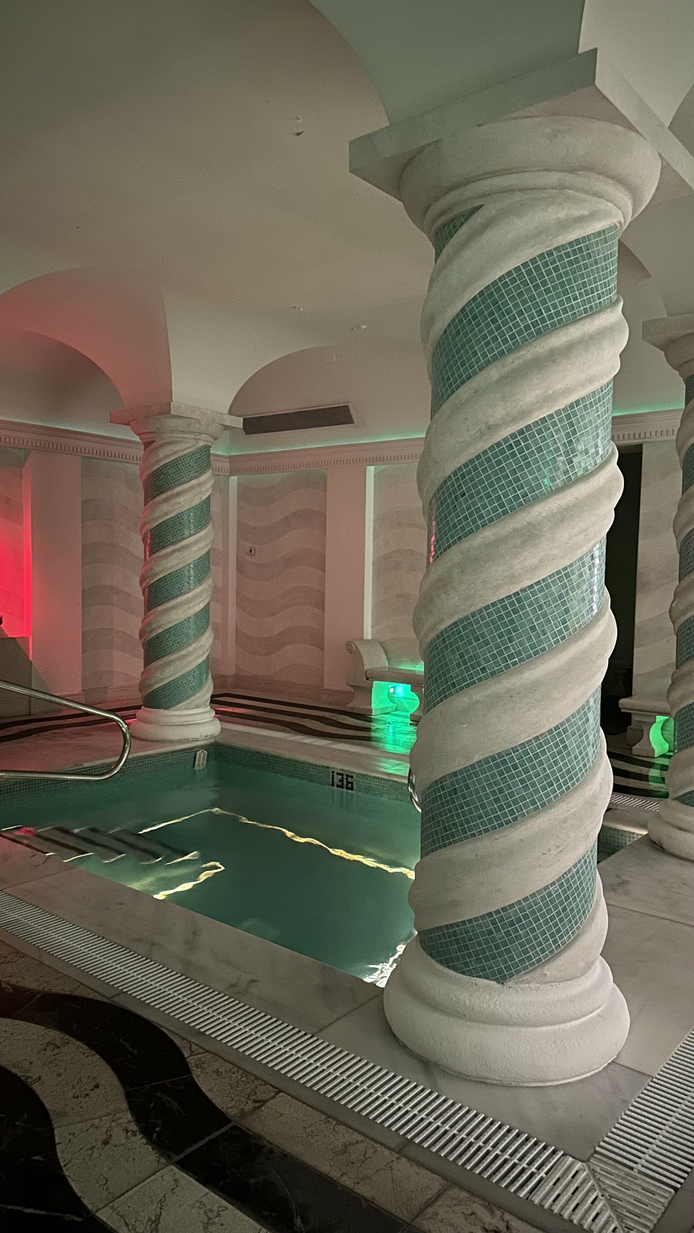 An indoor spa pool with mosaic-wrapped columns and arches in soft pink and green light