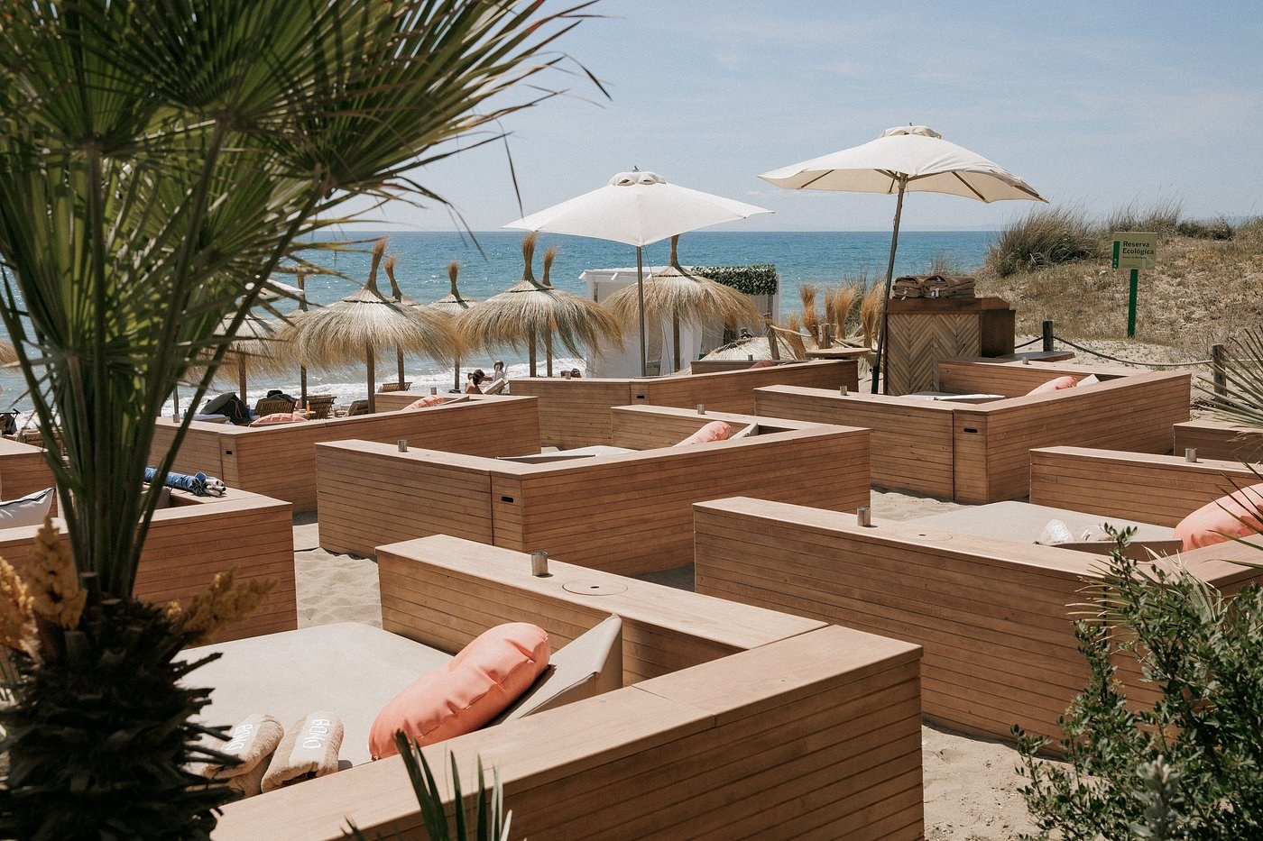 Timber banquette pods on the sand with thatched parasols and dune grass behind at Bono Beach, Marbella