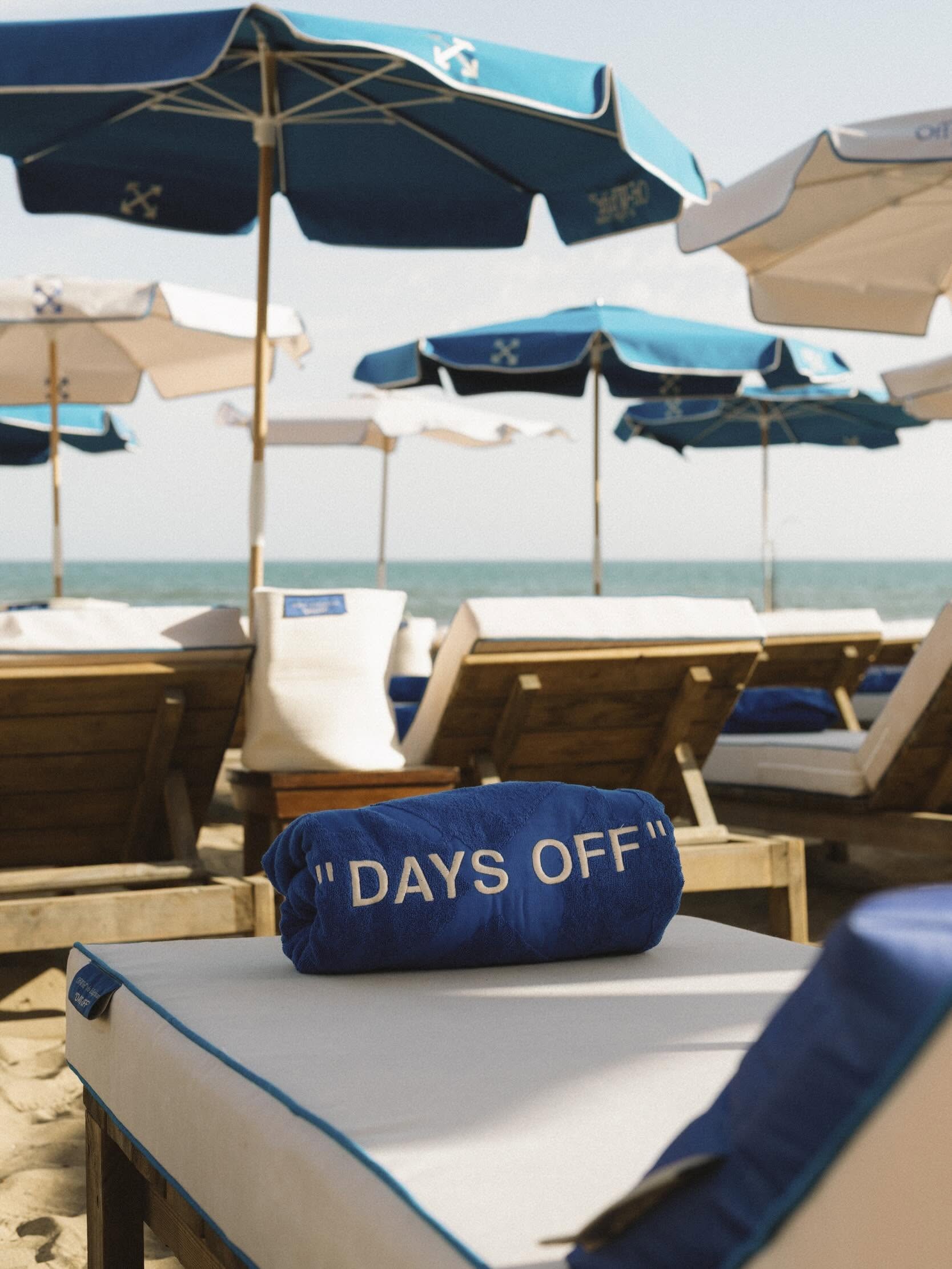 Navy and turquoise Off-White parasols with cross-arrow motifs above sunbeds, with a rolled blue towel reading Days Off