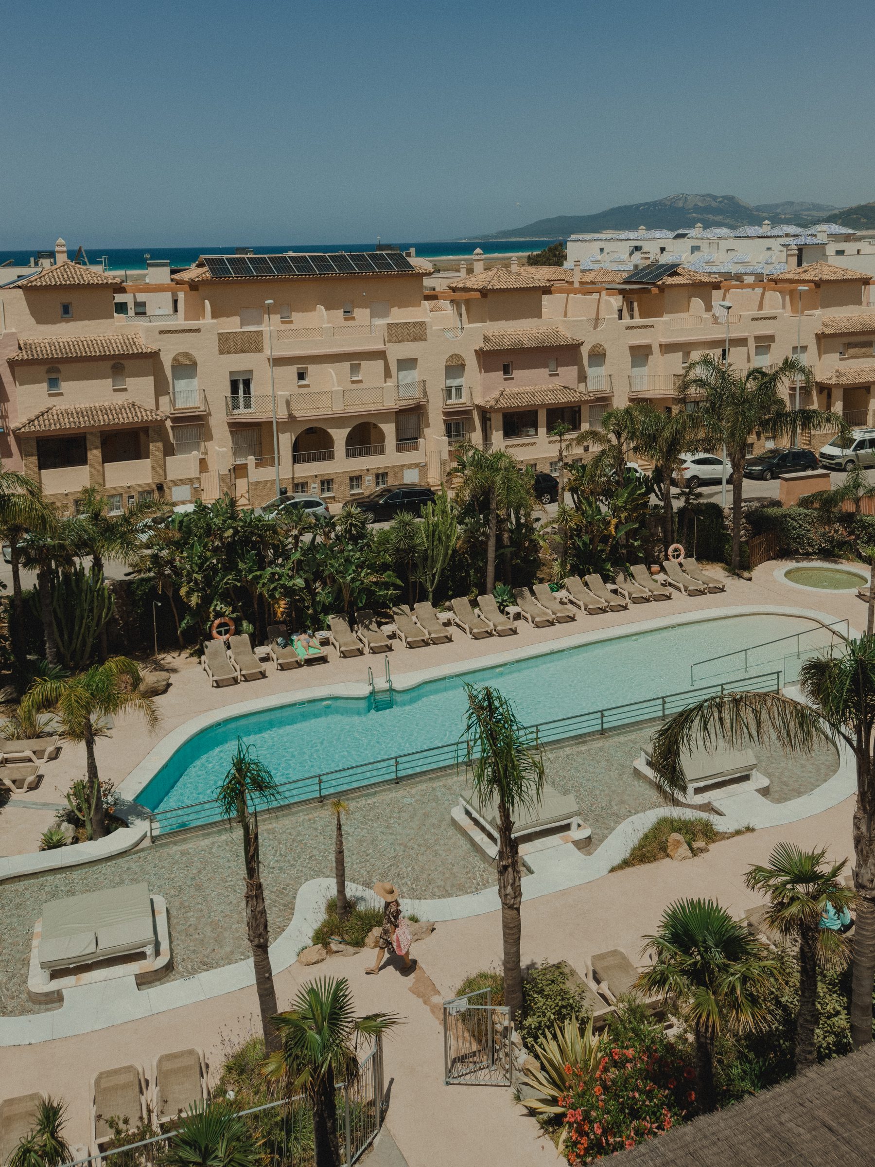 An aerial view of a curved hotel pool with palms, the sea and mountains beyond